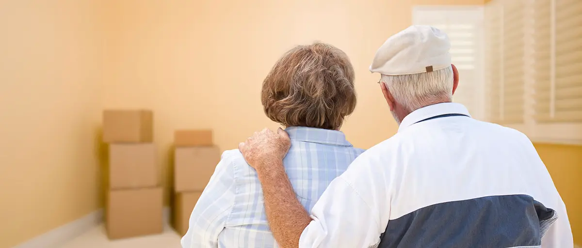 Retired couple standing arm in arm looking at moving boxes in an empty room before a downsizing move